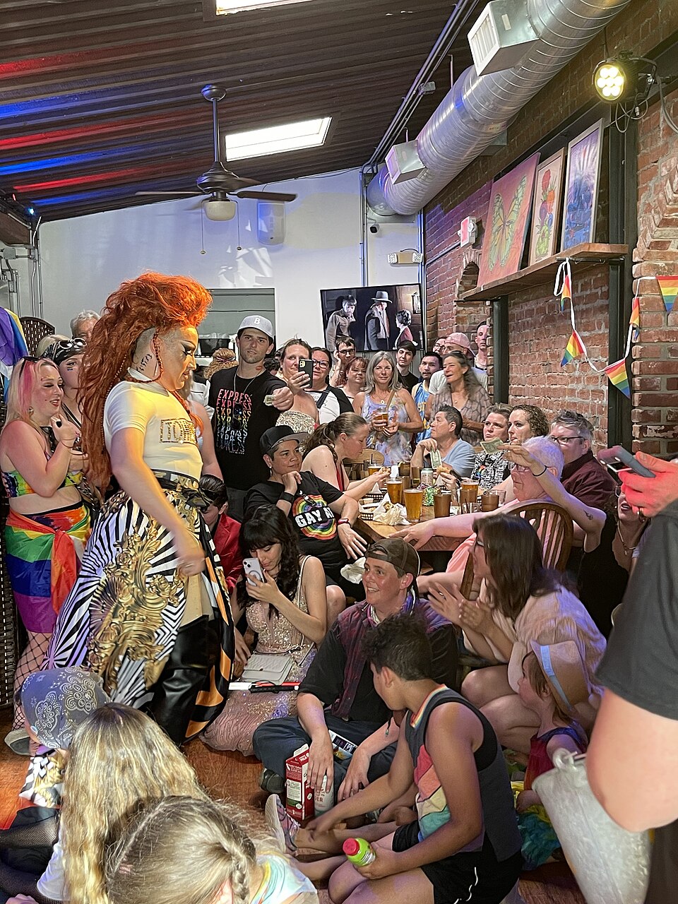 People of all genders gather in a festivally decorated bar for pride month. A drag performer marks the foreground.