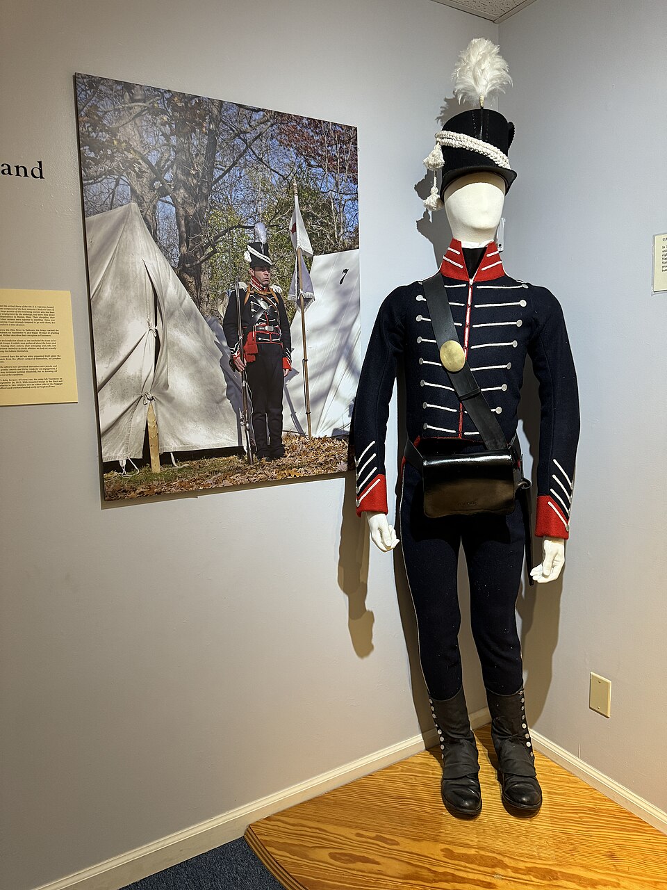 In a museum, a photograph featuring a historical soldier reenactor standing in a forest next to tents with a rifle, hangs next to a white faceless mannequin that sports the same soldier's outfit.