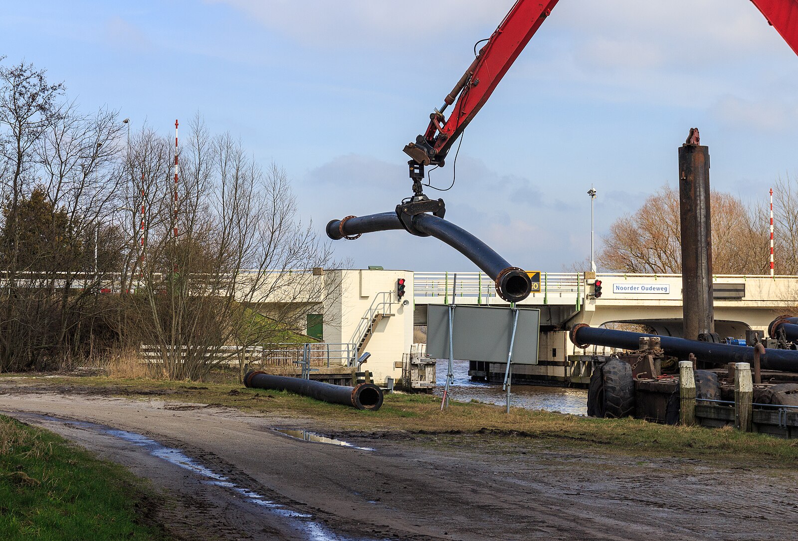 The arm of a large red crane towers over trees to pick up an enormous pipe.