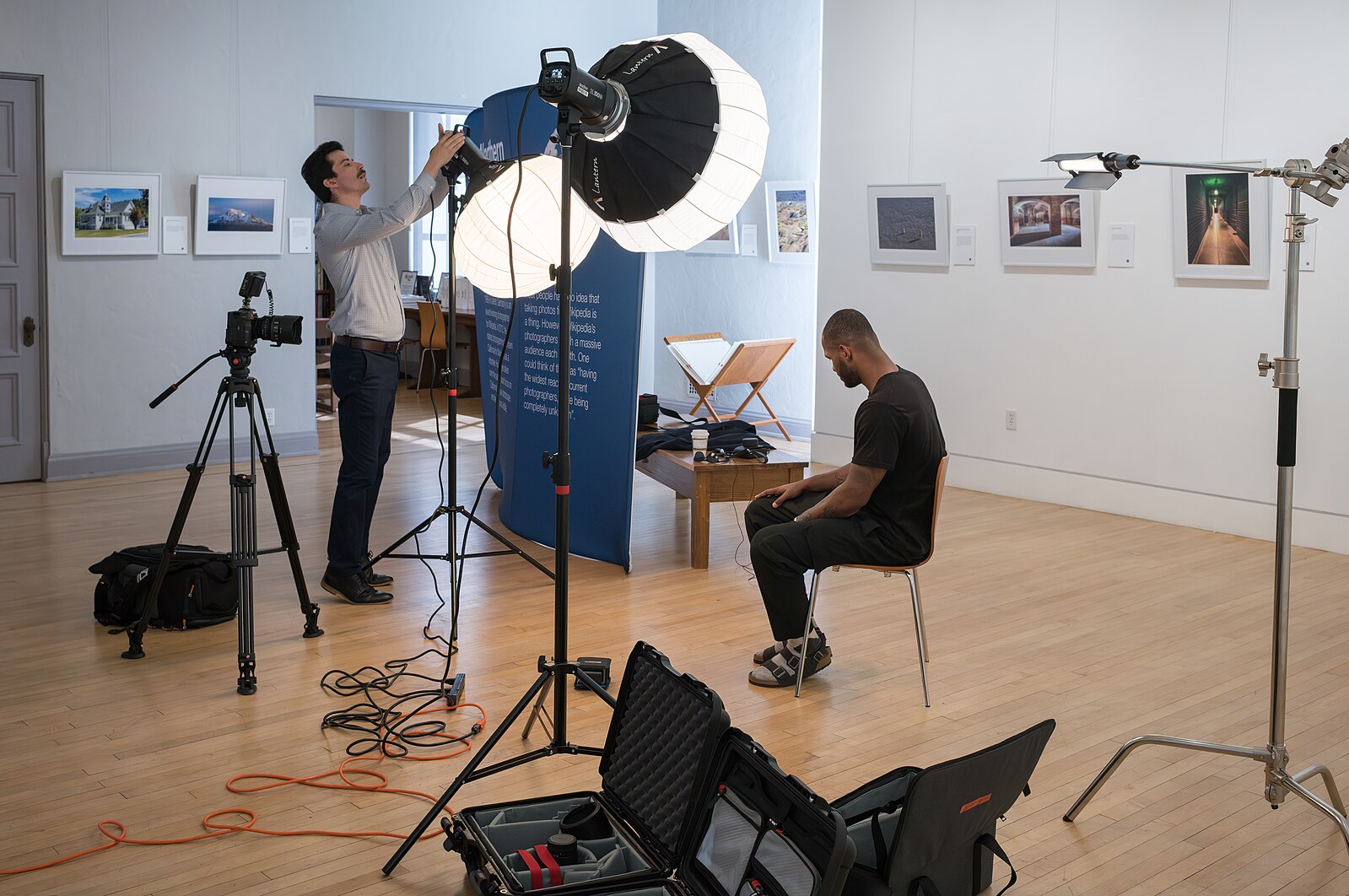 In an art gallery, one person sets up filming equipment while another person waits in a chair. The equipment includes many suitcases for item transport, three lights of two different types, the camera on a tripod, and sound equipemnt.