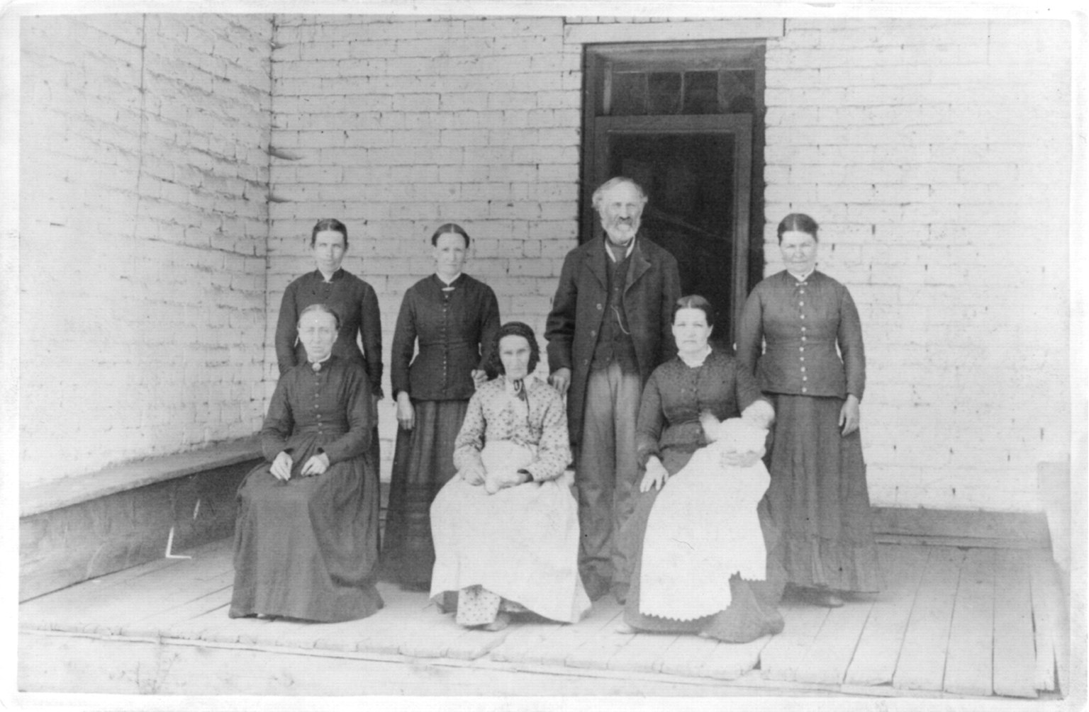 Old, black-and-white photograph of a man on a porch posing with family. A baby, and six women, the oldest is his mother. The others are neither his sisters nor his daughters.